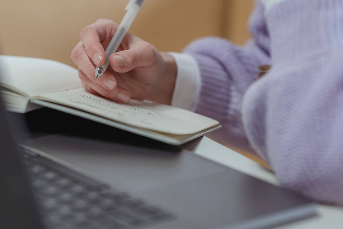 Hand schreibt mit einem Stift in ein aufgeschlagenes Notizbuch, das auf einem Laptop liegt