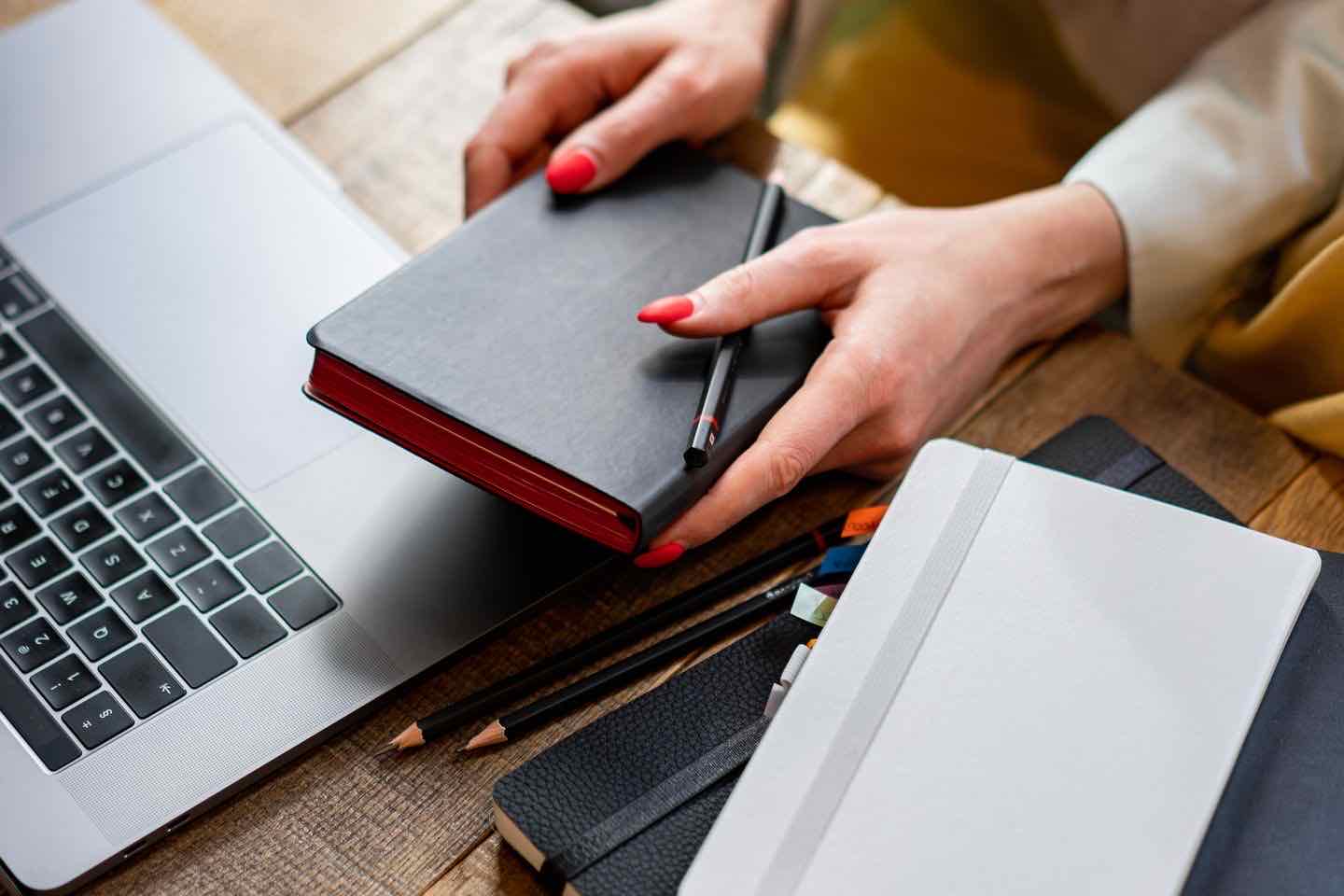 Hand hält ein Notizbuch mit Stift neben einem aufgeklappten Laptop
